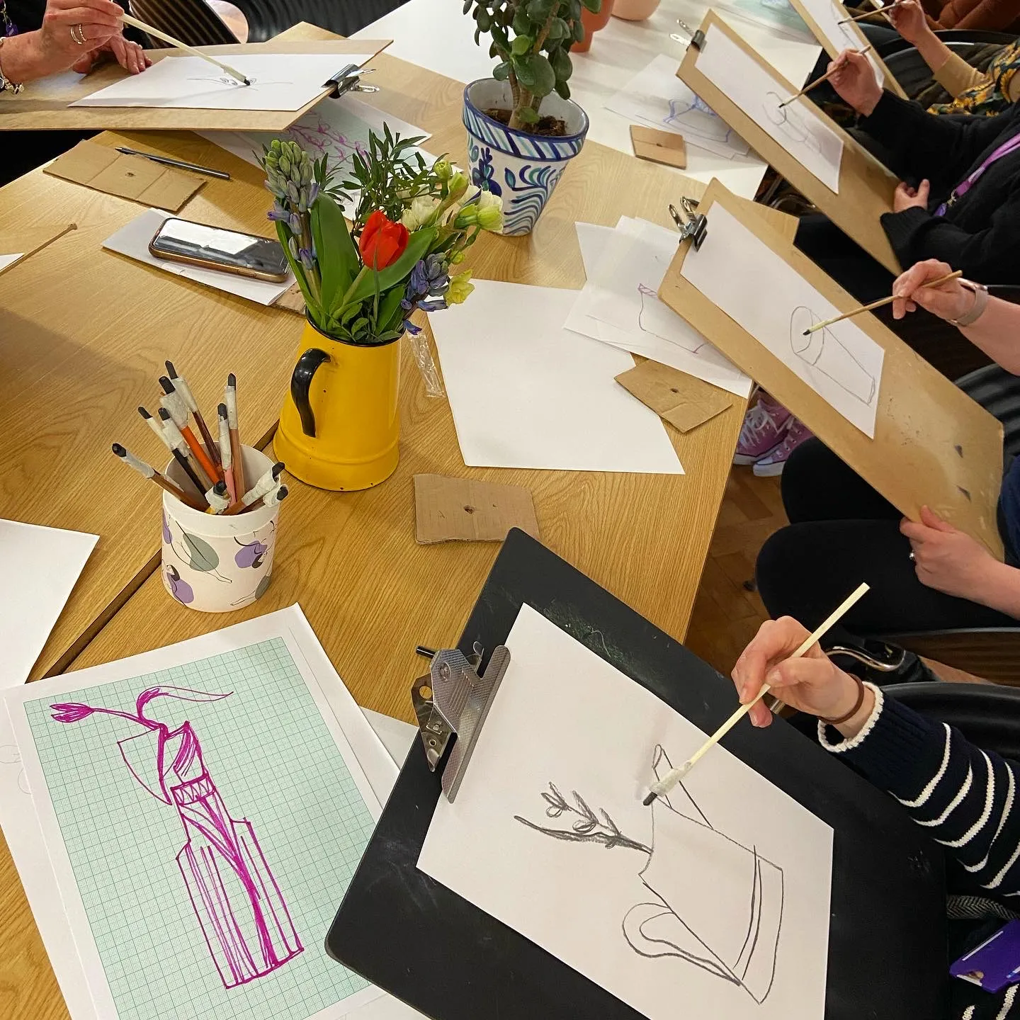 Adults sketching flowers in a vase during a class at Art Club, brushes standing in a yellow jug